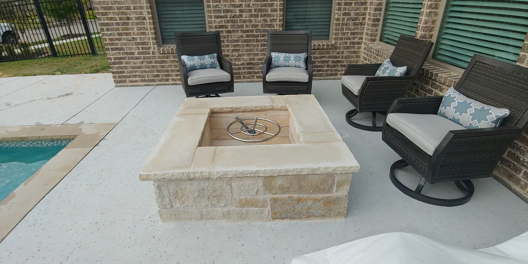 Square masonry fire pit and chairs on a backyard patio beside a pool